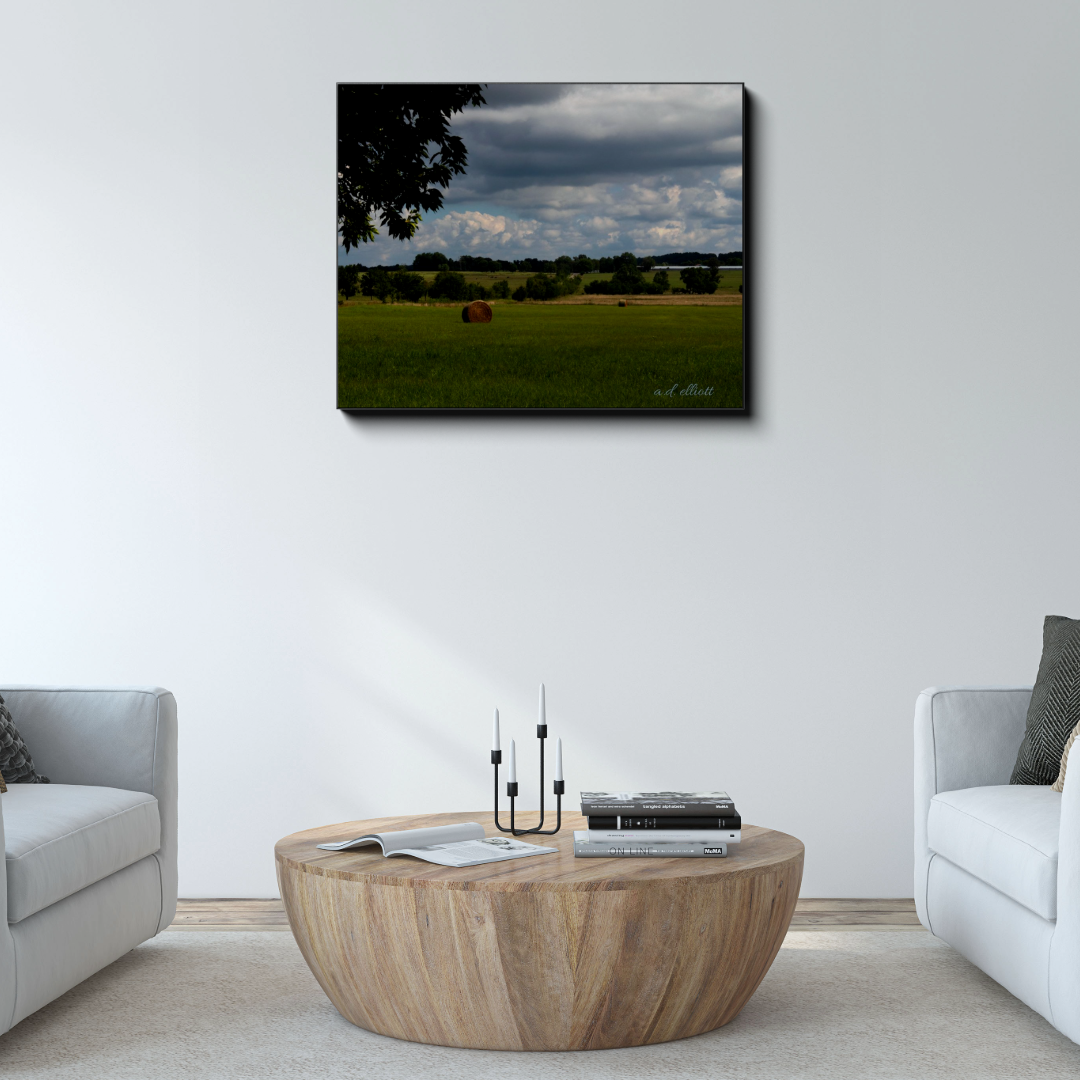 Arkansas Hayfield canvas photograph displayed above a round wooden coffee table between two light-colored chairs in a bright living room.