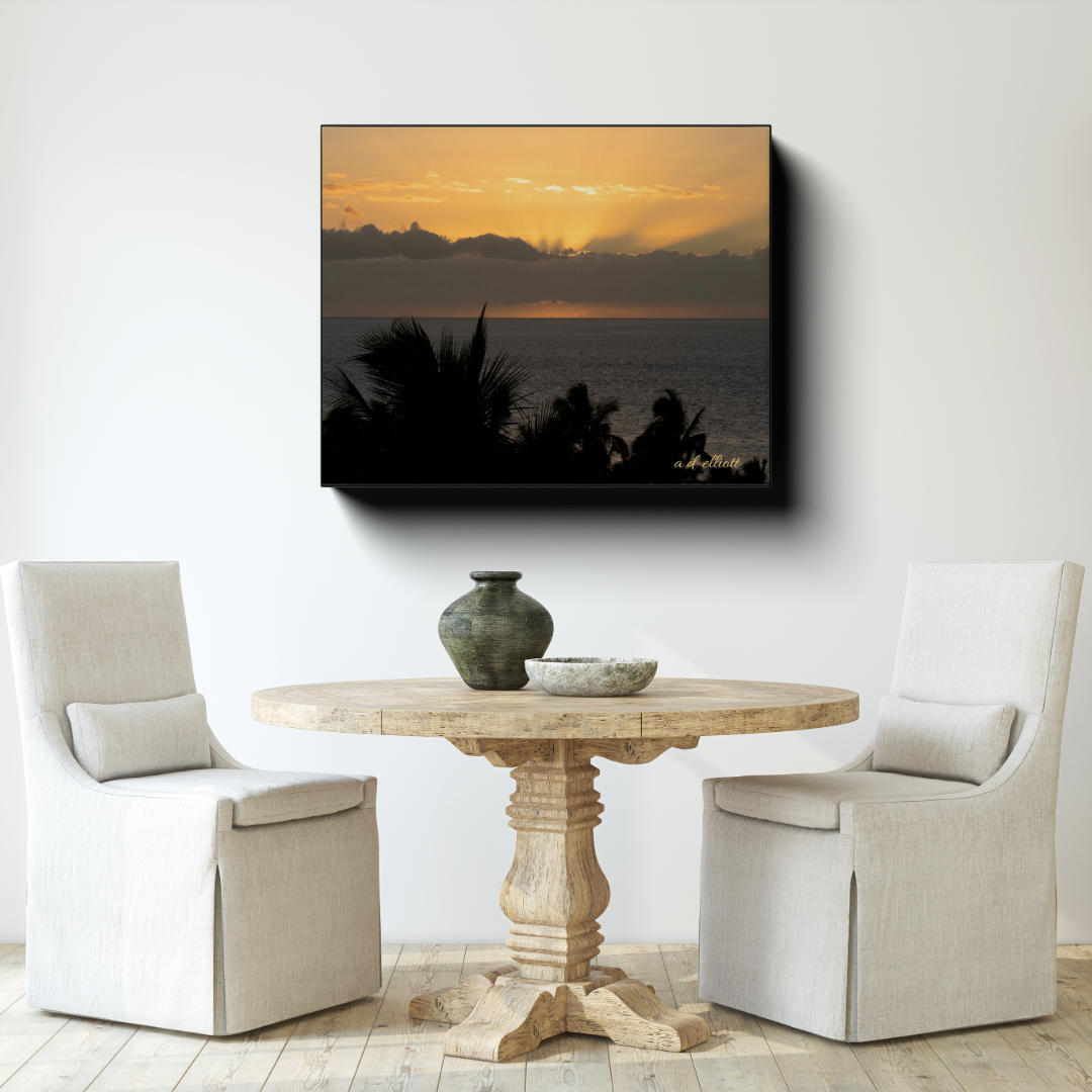 Gallery-wrapped canvas featuring a golden Maui sunset over the Pacific Ocean displayed above a round wooden table in a bright neutral dining area.