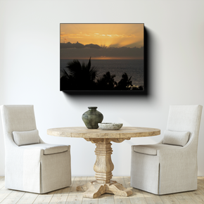 Gallery-wrapped canvas featuring a golden Maui sunset over the Pacific Ocean displayed above a round wooden table in a bright neutral dining area.