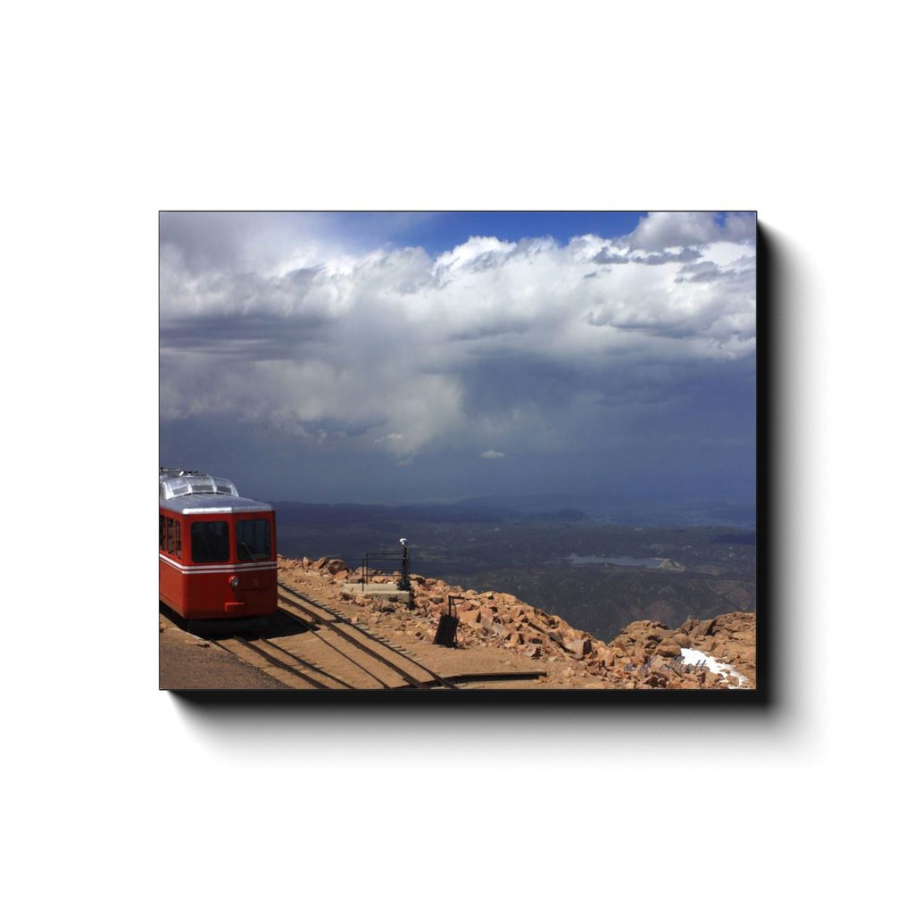 Gallery-wrapped canvas print featuring the red Pikes Peak cog train, rocky mountain tracks, distant valleys, and dramatic clouds.