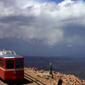 Gallery-wrapped canvas print featuring the red Pikes Peak cog train, rocky mountain tracks, distant valleys, and dramatic clouds.